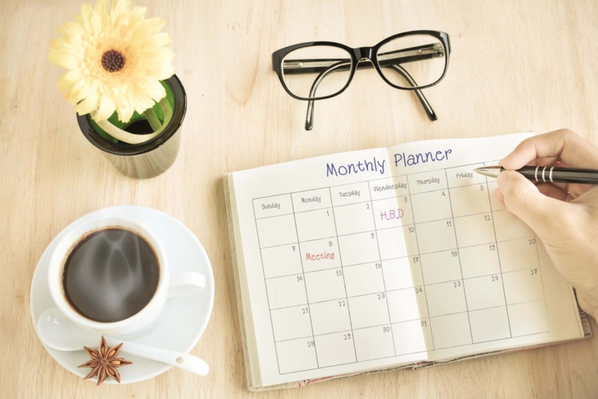 Hand filling out a monthly planner next to a cup of coffee and a flower